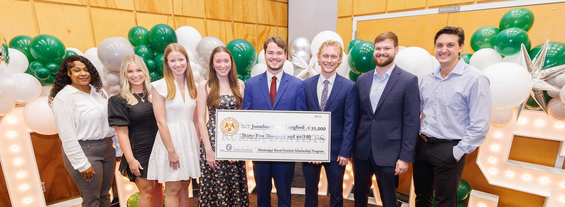 Group standing indoors holding an oversized scholarship check in front of green, white, and silver balloon decorations.